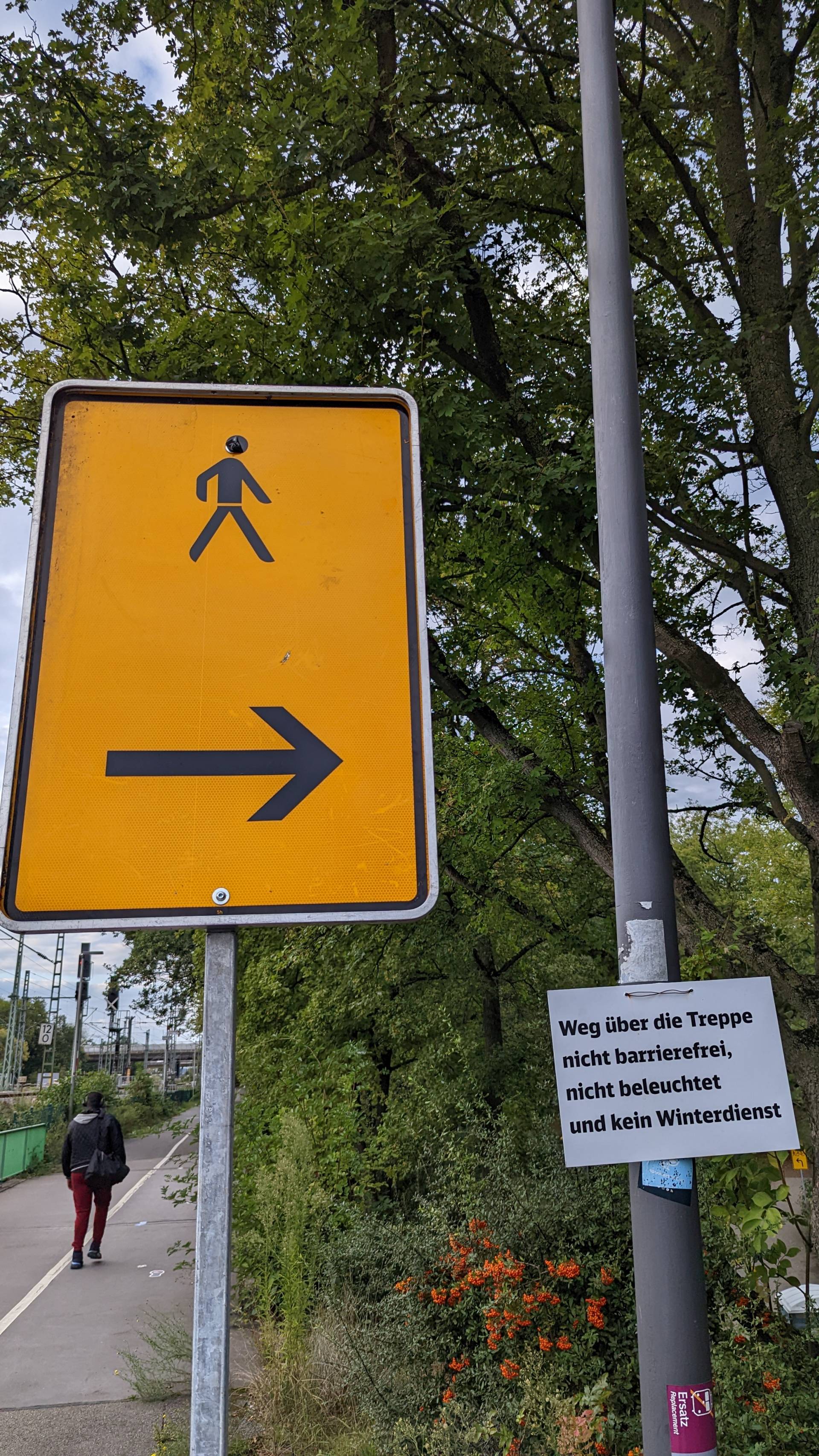 Ein orangenes Schild, dass einen Fußgänger zeigt und den Weg mit einem Pfeil nach rechts auf den Weg rechts weißt. Daneben klebt ein laminiertes Blatt auf dem steht: "Weg über die Treppe nicht barrierefrei, nicht beleuchtet und kein Winterdienst&