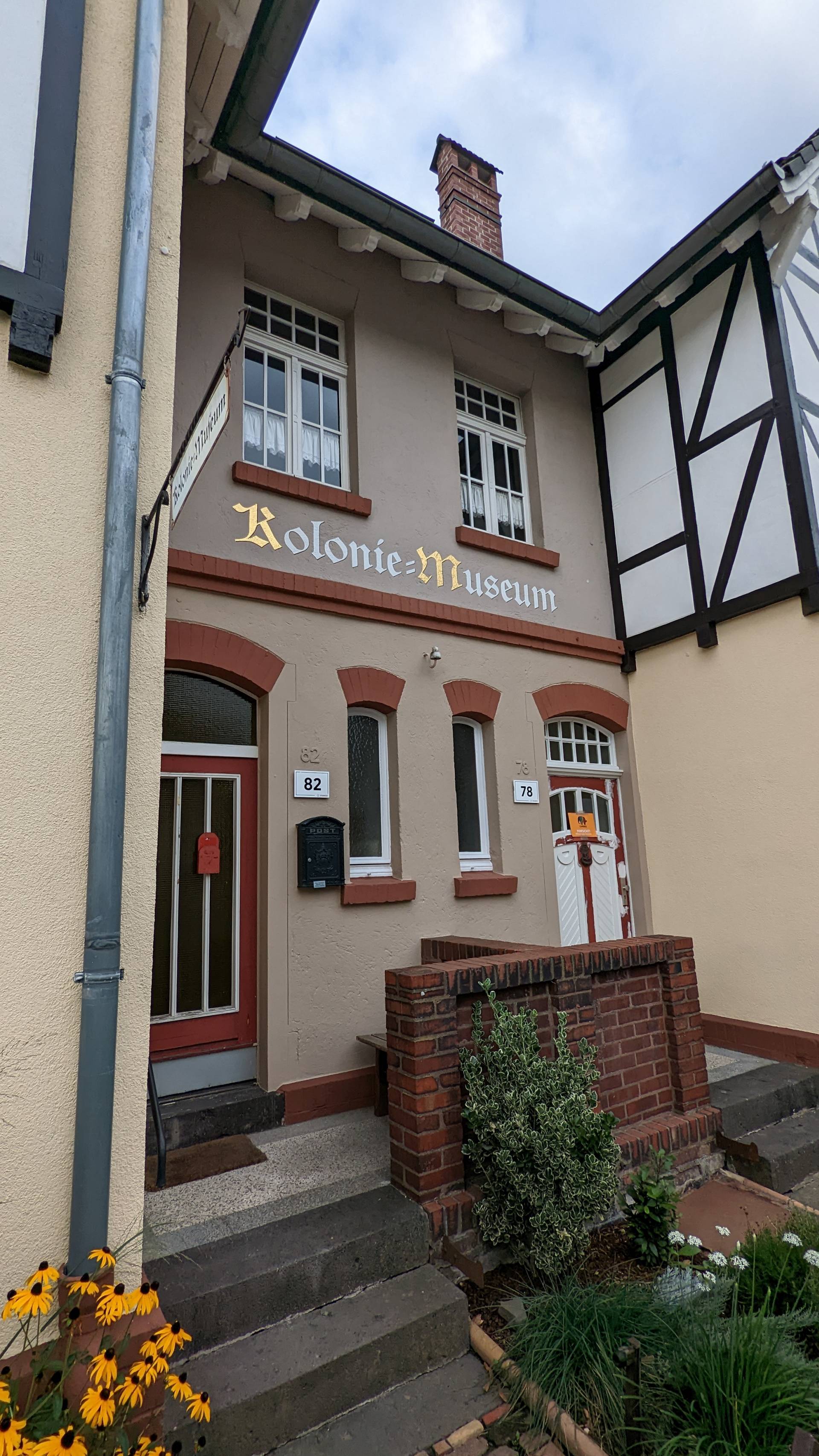 Der Eingang eines Reihendoppelhauses. In der Mitte zwischen den beiden Hauseingängen steht in großer Schrift "Kolonie-Museum". Die Anfangsbuchstaben der Schrift sind golden, die anderen Buchstaben weiß.