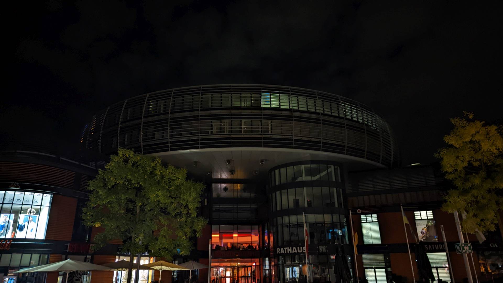 Die Rathaus-Galerie und das Rathaus in der Innenstadt. Der Himmel ist dunkel. Einige Geschäfte haben Licht an und beleuchten die Schaufenster. Im Rathaus brennt noch ein leichtes Licht in einem Teil, der Rest ist im Dunkeln.