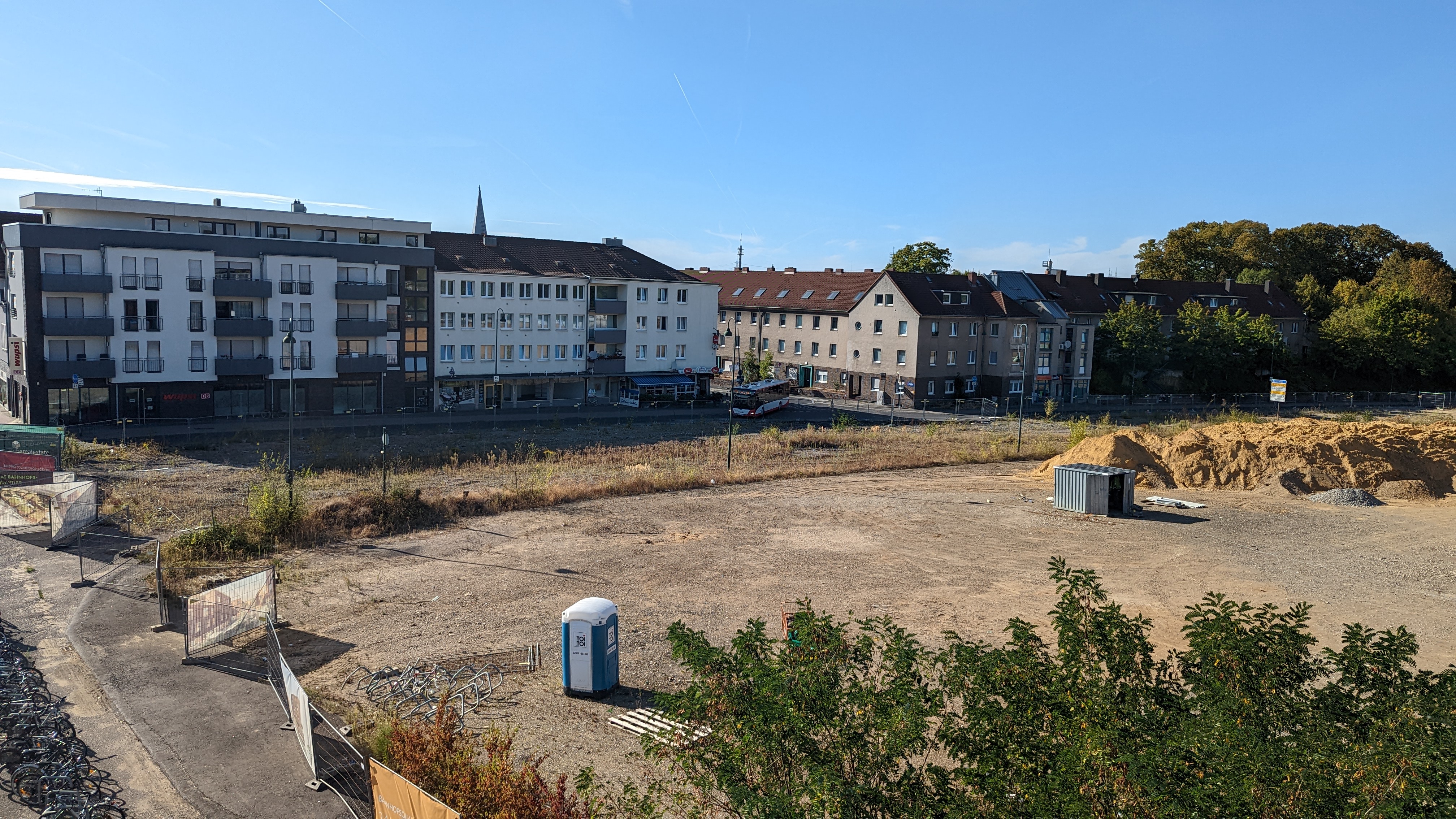 Aufnahme von der Bahnhofsbrücke runter zum alten Busbahnhof in Opladen. Der alte Busbahnhof ist entfernt und zu sehen ist eine Baustelle mit Kies und Sandhaufen. Die Baustelle ist von Bauzäunen umzäunt und somit abgesperrt.